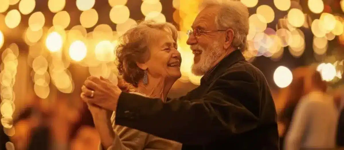 Casal de idosos dançando juntos, rodeados por luzes festivas e sorrindo, em uma celebração social.