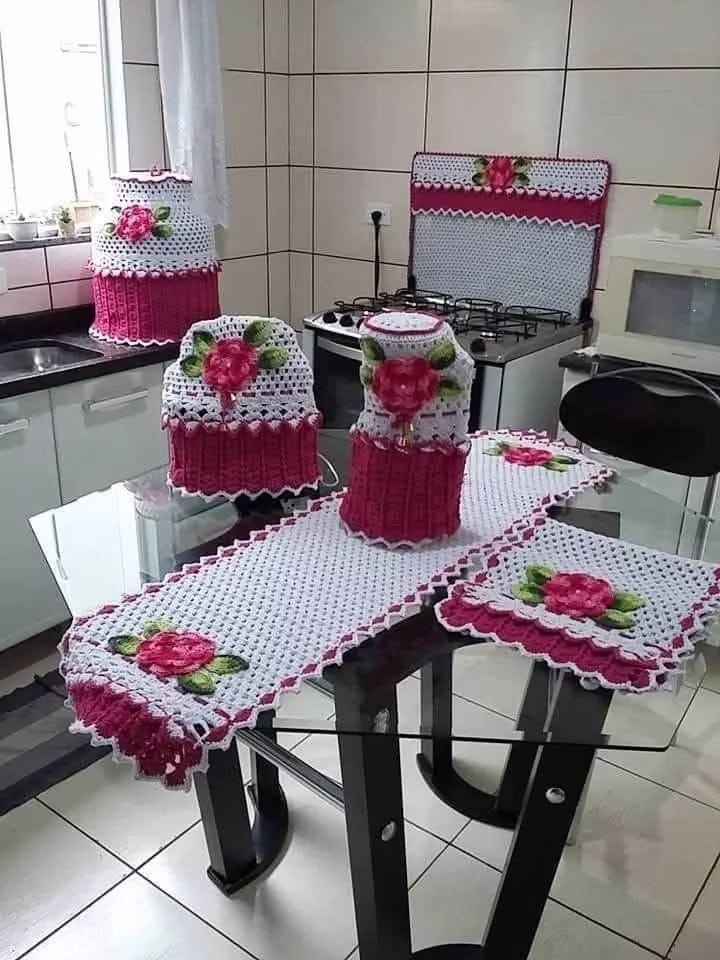 Conjunto de utensílios de crochê na cozinha, incluindo toalha de mesa, capas para potes e forno, com detalhes em rosa e branco, trazendo charme ao ambiente.
