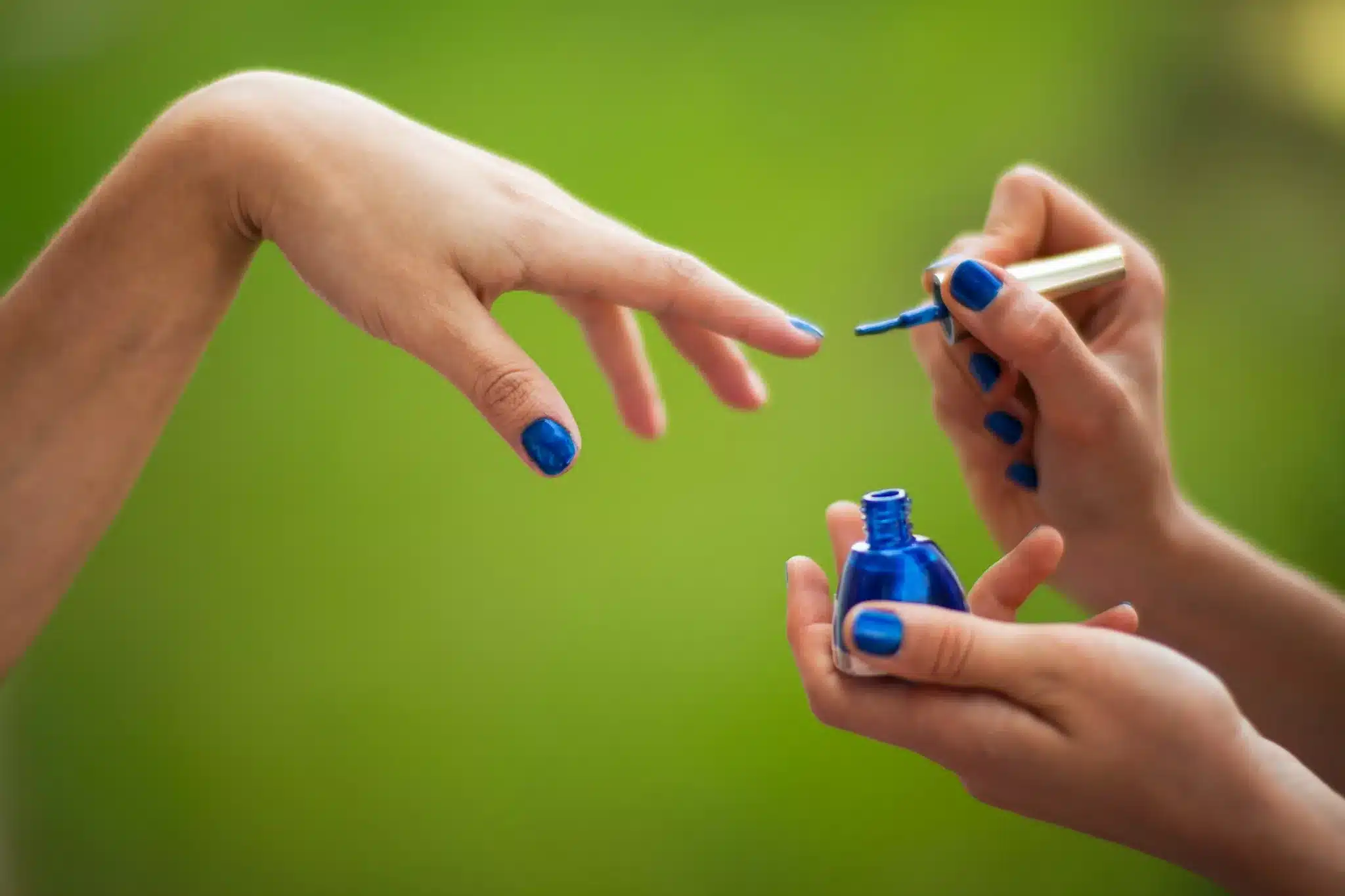 Mulher aplicando esmalte vegano azul em outra mulher em ambiente natural.