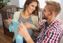 Casal feliz esperando o nascimento do filho, segurando uma roupa de bebê, com o berço ao fundo, celebrando a chegada de um novo membro na família.