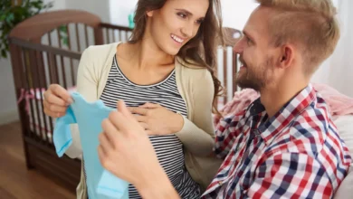 Casal feliz esperando o nascimento do filho, segurando uma roupa de bebê, com o berço ao fundo, celebrando a chegada de um novo membro na família.
