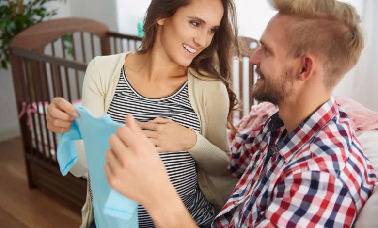 Casal feliz esperando o nascimento do filho, segurando uma roupa de bebê, com o berço ao fundo, celebrando a chegada de um novo membro na família.