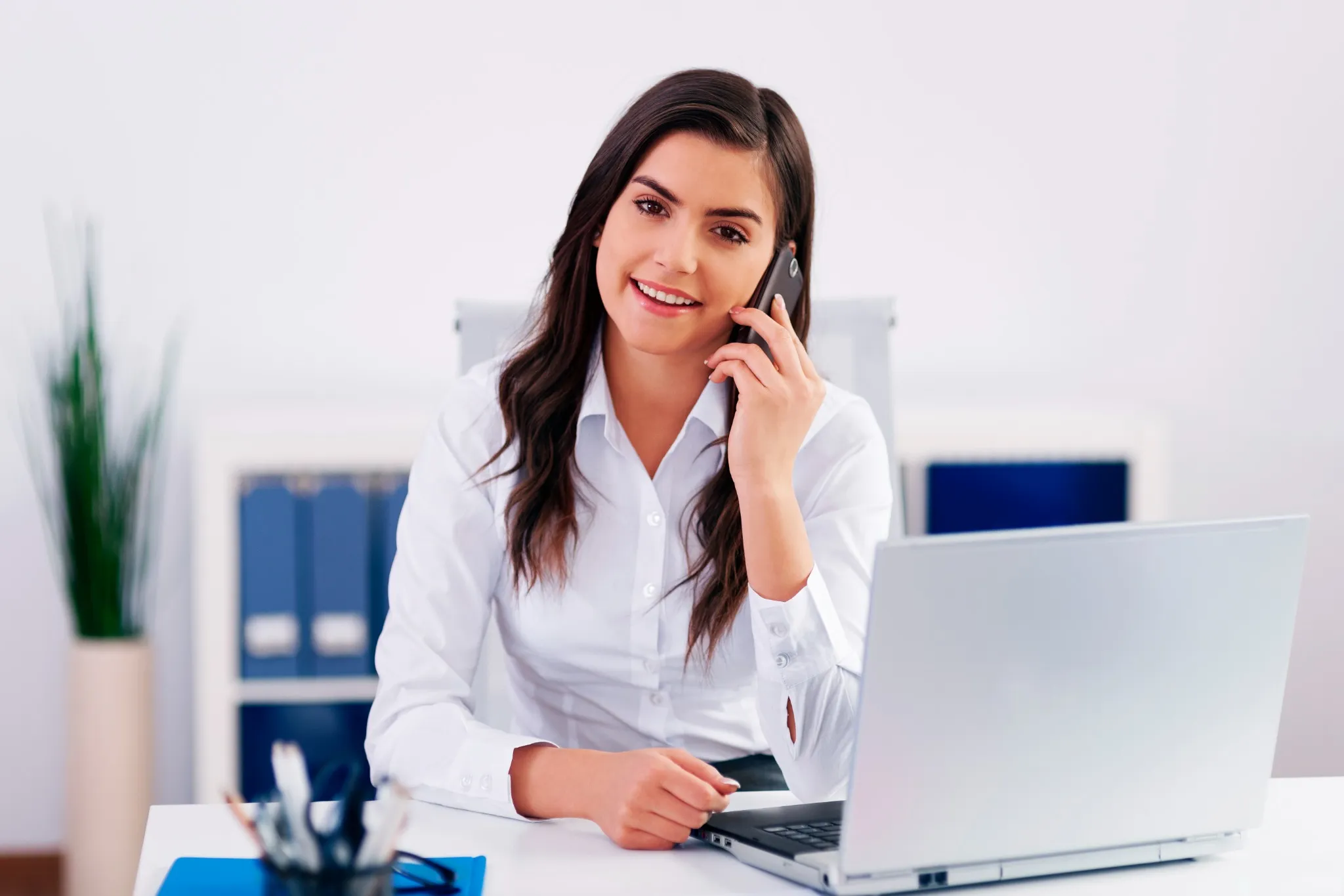 Executiva de atendimento ao cliente no escritório, sorrindo enquanto fala ao telefone com um laptop aberto à sua frente.