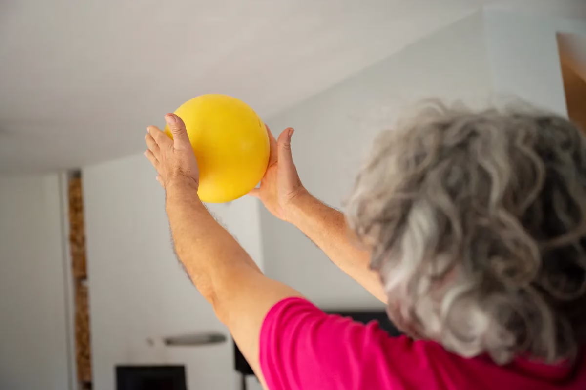 Idosa segurando bola amarela durante exercício em casa.