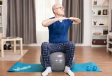 Idoso praticando exercícios leves sentado em uma bola suíça, realizando alongamento dentro de sua casa.