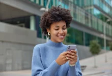 Jovem sorridente com cabelo cacheado, segurando o celular, transmitindo alegria e boas vibrações para um fim de semana incrível.