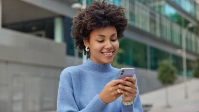 Jovem sorridente com cabelo cacheado, segurando o celular, transmitindo alegria e boas vibrações para um fim de semana incrível.
