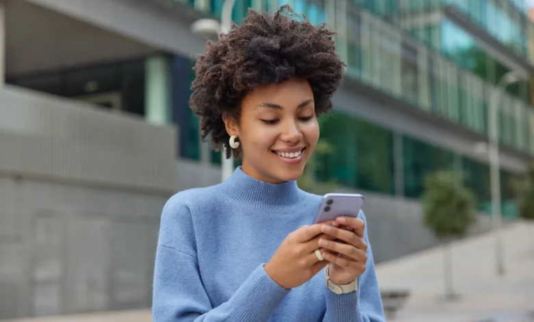 Jovem sorridente com cabelo cacheado, segurando o celular, transmitindo alegria e boas vibrações para um fim de semana incrível.