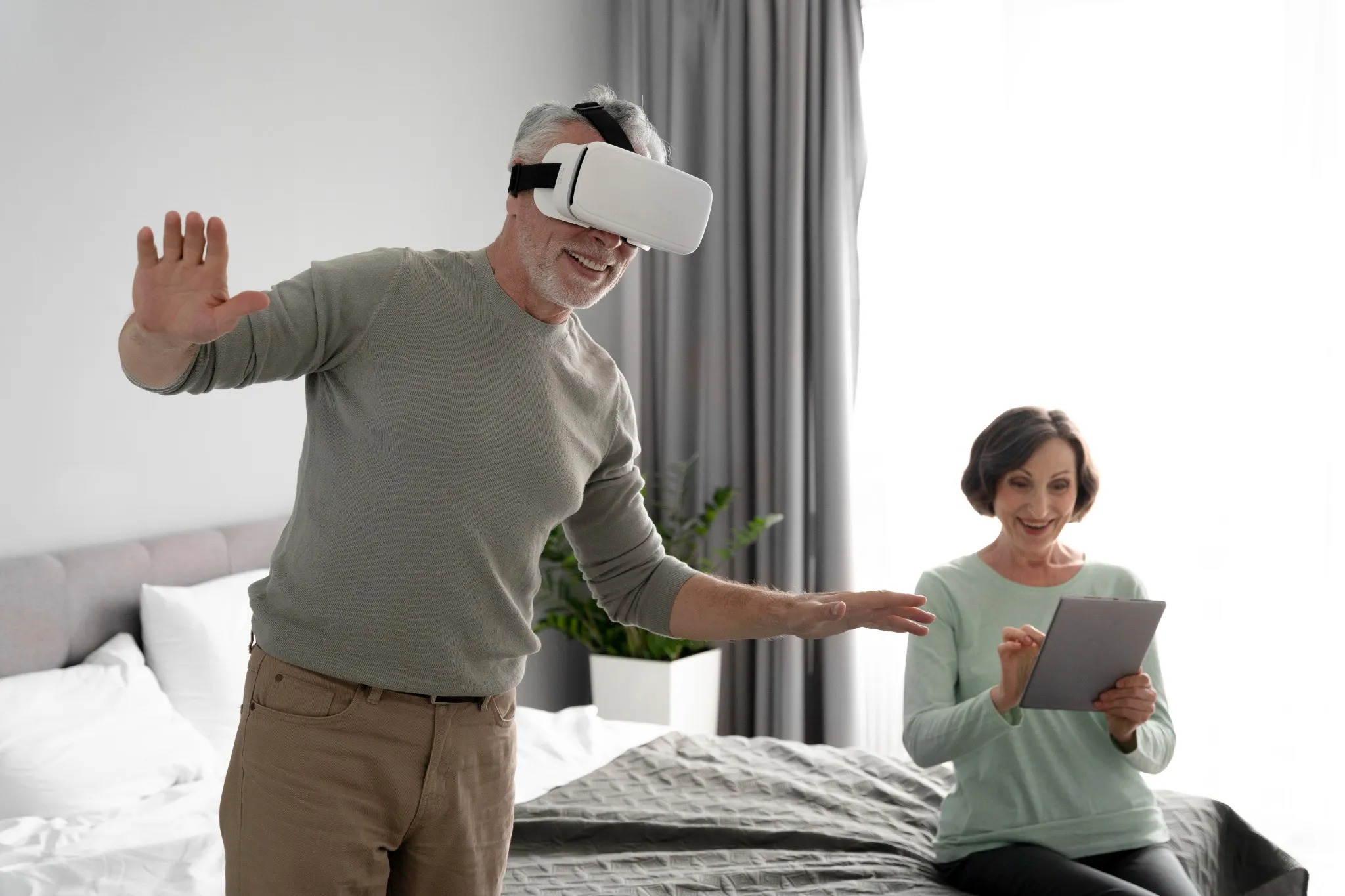 Pessoa idosa jogando com óculos de realidade virtual enquanto outra assiste, usando um tablet.