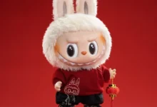 Boneco fofinho com capô de coelho, roupa vermelha e tênis, segurando lanterna de decoração.