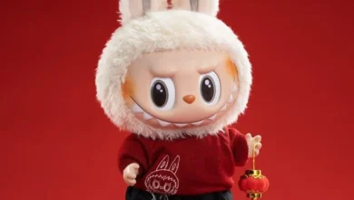 Boneco fofinho com capô de coelho, roupa vermelha e tênis, segurando lanterna de decoração.