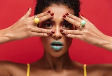 Mulher com maquiagem ousada e anéis coloridos, destacando as unhas vermelhas e o fundo vermelho vibrante.