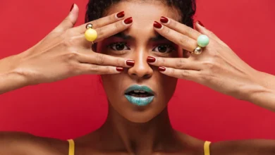 Mulher com maquiagem ousada e anéis coloridos, destacando as unhas vermelhas e o fundo vermelho vibrante.