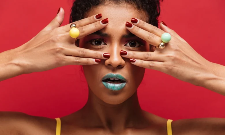 Mulher com maquiagem ousada e anéis coloridos, destacando as unhas vermelhas e o fundo vermelho vibrante.