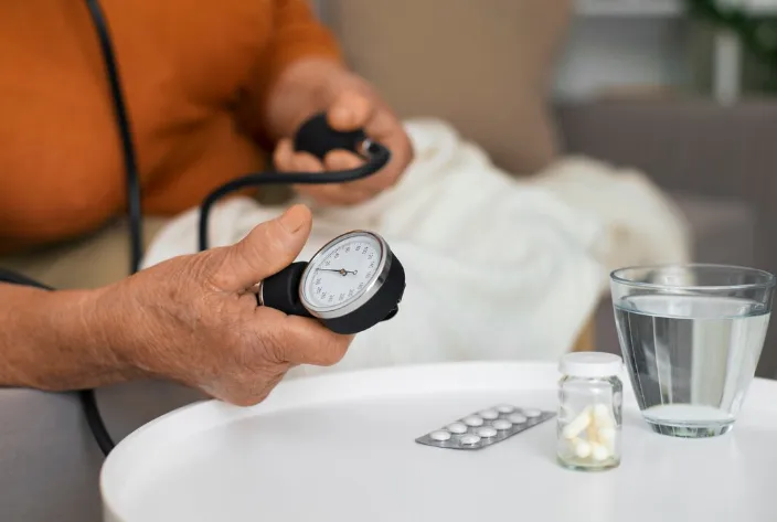 Idoso monitorando sua pressão arterial enquanto ao lado há medicamentos e água, simbolizando cuidados de saúde na terceira idade.