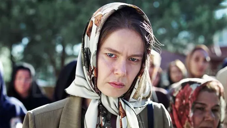 Mulher com expressão séria usando lenço floral na cabeça em cena de drama turco.