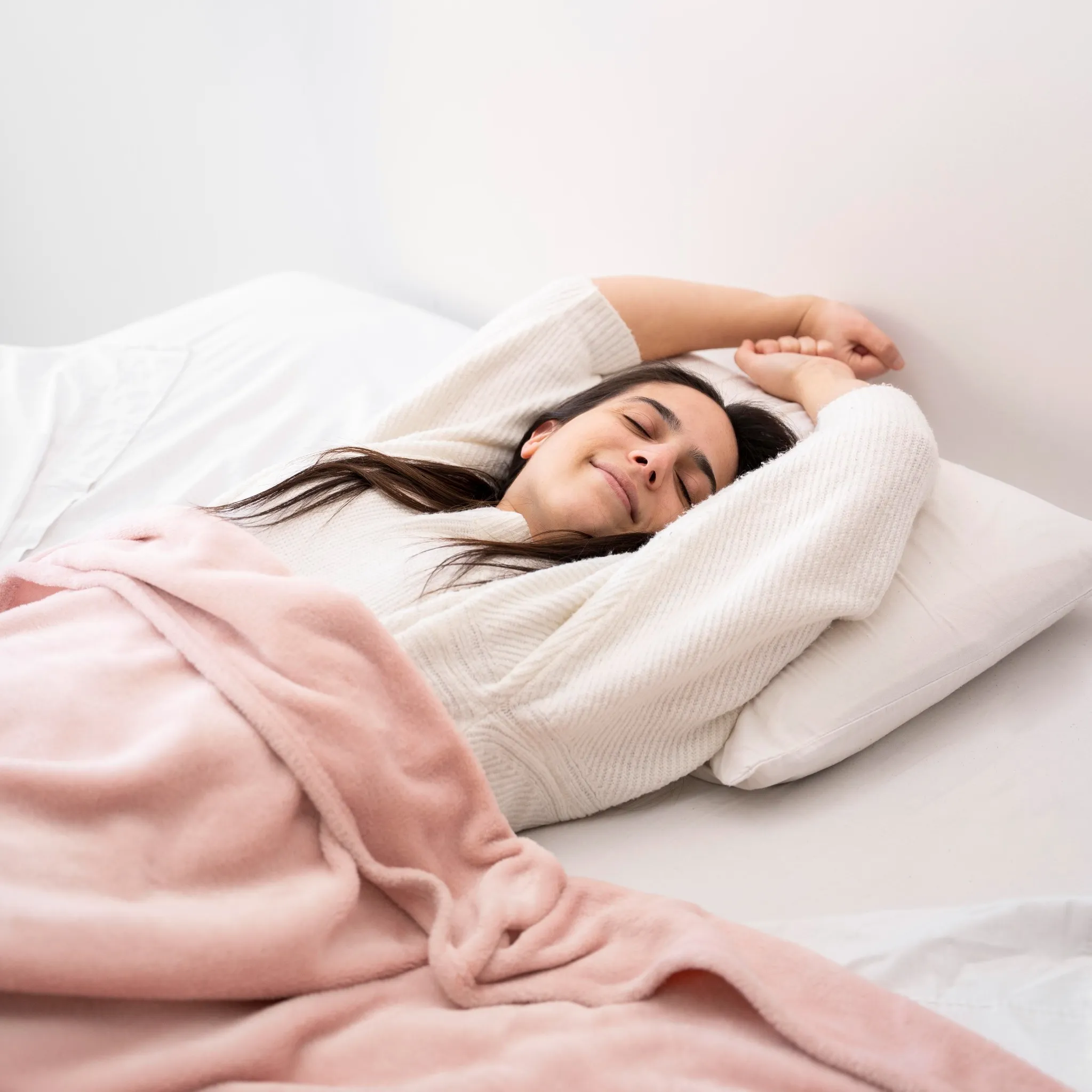 Mulher deitada na cama sorrindo, com os braços esticados para cima, demonstrando relaxamento após uma boa noite de sono.