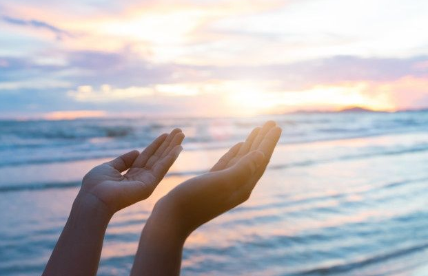 Mãos abertas ao entardecer na praia, com luz suave, simbolizando uma mensagem de paz e bênçãos para o mês de julho.