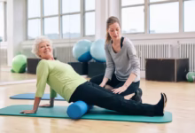 Idosa praticando pilates com auxílio de profissional em estúdio.