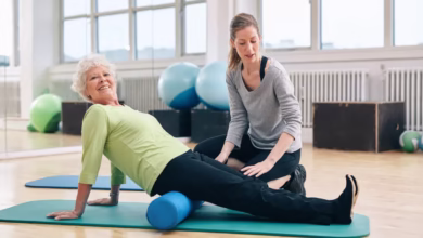 Idosa praticando pilates com auxílio de profissional em estúdio.