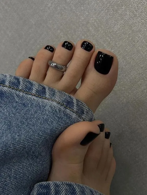 Unhas dos pés pintadas de preto, com anel prateado e jeans desbotados