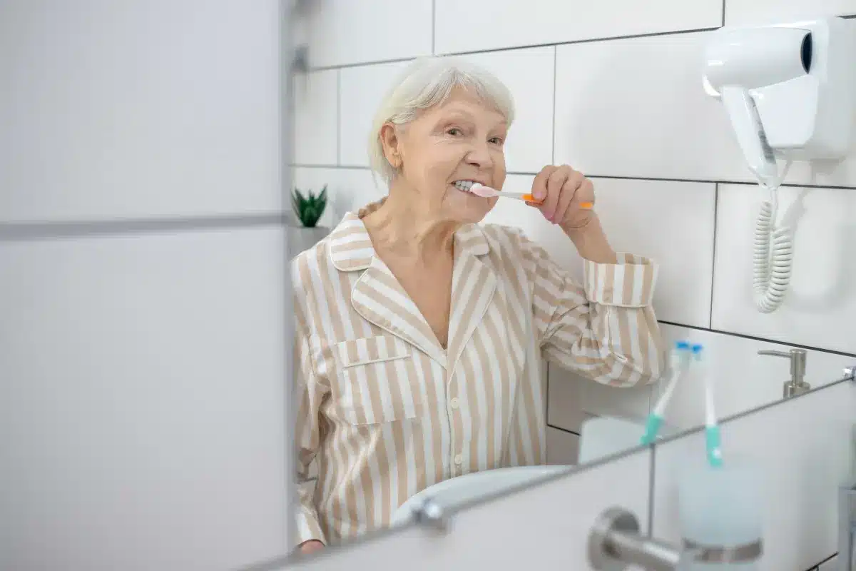 Idosa escovando os dentes em frente ao espelho do banheiro.