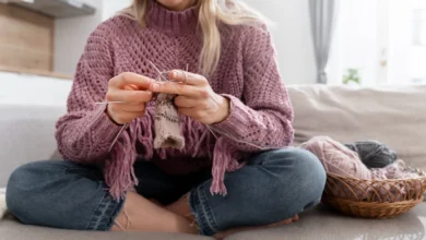 Mulher jovem tricotando tranquilamente em sofá, com fios coloridos ao lado.