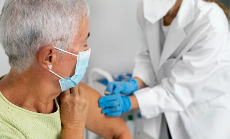Idosa recebendo vacina contra doenças em uma clínica de saúde com a ajuda de um profissional médico usando máscara e luvas.