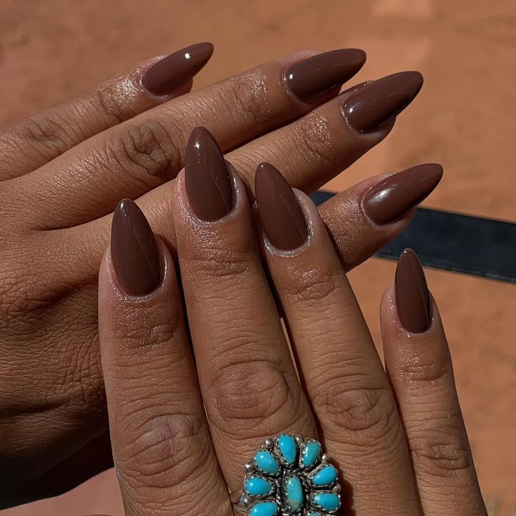 A imagem de uma mão com as unhas longas e pontiagudas, pintadas de um tom de marrom escuro, com um anel turquesa no dedo.