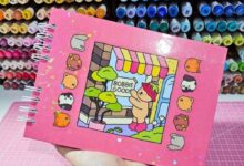 Caderno com ilustração de canetinhas Bobbie Goods e personagem com fantasia de morango, ilustrando um cenário de loja.