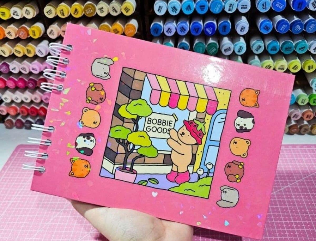 Caderno com ilustração de canetinhas Bobbie Goods e personagem com fantasia de morango, ilustrando um cenário de loja.