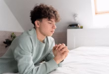 Jovem em oração ajoelhado ao lado da cama, simbolizando uma noite de paz e reflexão.