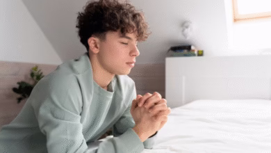 Jovem em oração ajoelhado ao lado da cama, simbolizando uma noite de paz e reflexão.