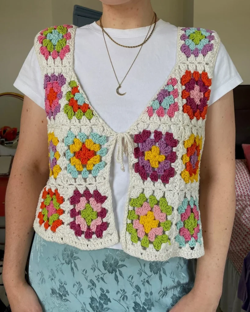 Imagem de um colete feito em crochê com quadrados granny coloridos, como um padrão vibrante de flores, com fundo claro e laço frontal.