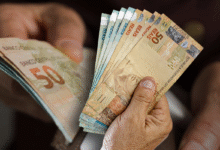 Uma mão segurando o cartão do Bolsa Família e diversas notas de R$ 100, representando a transferência de renda do governo para famílias brasileiras.