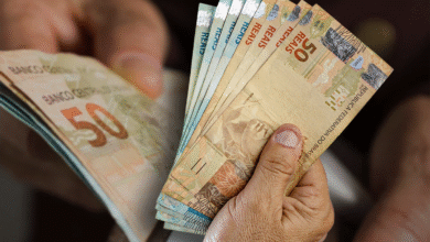 Uma mão segurando o cartão do Bolsa Família e diversas notas de R$ 100, representando a transferência de renda do governo para famílias brasileiras.