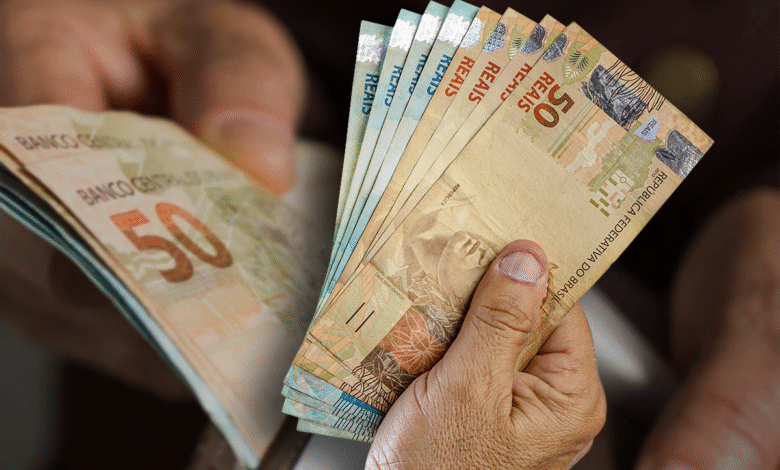 Uma mão segurando o cartão do Bolsa Família e diversas notas de R$ 100, representando a transferência de renda do governo para famílias brasileiras.