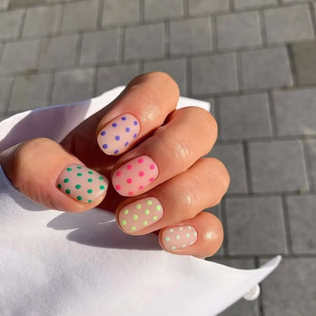 Unhas curtas com esmalte em tons pastel e bolinhas coloridas.