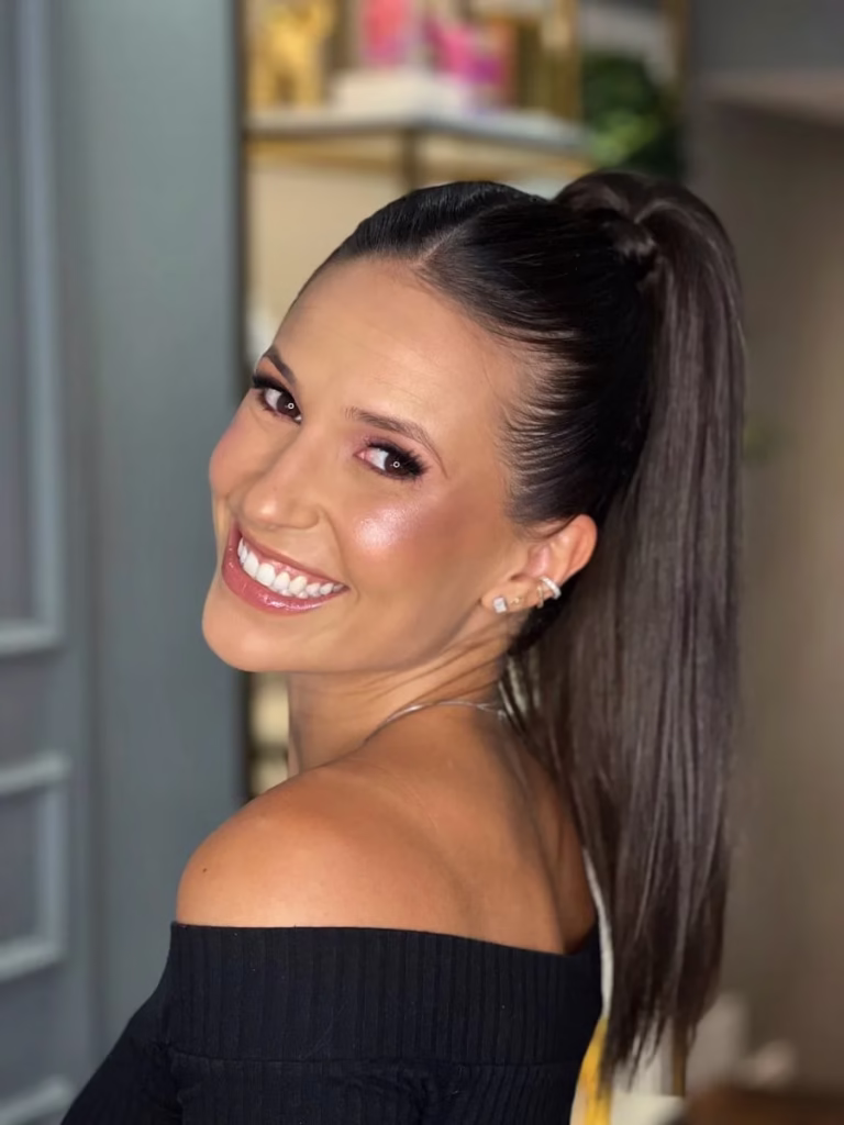 Mulher sorrindo com rabo de cavalo partido ao meio e volume, usando uma blusa preta de ombro a ombro.