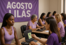 Mulheres em uma sala com banner escrito 'Agosto Lilás', utilizando computadores e realizando atendimento