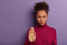 Mulher com uma expressão séria e mão levantada, demonstrando rejeição ou pedido para parar, vestindo um suéter roxo em fundo lilás.