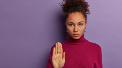 Mulher com uma expressão séria e mão levantada, demonstrando rejeição ou pedido para parar, vestindo um suéter roxo em fundo lilás.