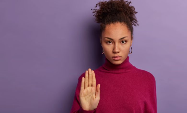 Mulher com uma expressão séria e mão levantada, demonstrando rejeição ou pedido para parar, vestindo um suéter roxo em fundo lilás.
