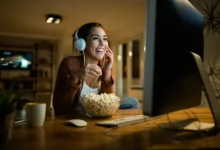 Mulher sorridente com fones de ouvido assiste a novela online enquanto come pipoca.