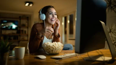 Mulher sorridente com fones de ouvido assiste a novela online enquanto come pipoca.