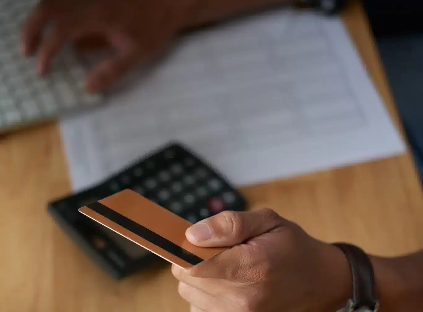 Homem segurando cartão de crédito com foco em mãos, teclado e calculadora ao fundo, simbolizando a gestão de pagamentos e faturas.