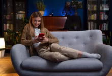 Mulher sorrindo enquanto assiste algo no celular em uma noite tranquila, com uma iluminação aconchegante no fundo.