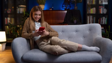 Mulher sorrindo enquanto assiste algo no celular em uma noite tranquila, com uma iluminação aconchegante no fundo.