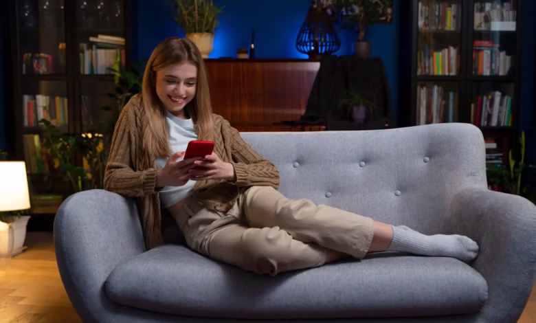 Mulher sorrindo enquanto assiste algo no celular em uma noite tranquila, com uma iluminação aconchegante no fundo.