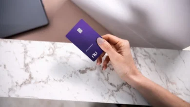 Cartão Nubank ultravioleta sendo segurado por uma pessoa sobre uma mesa de mármore, em um cenário minimalista.
